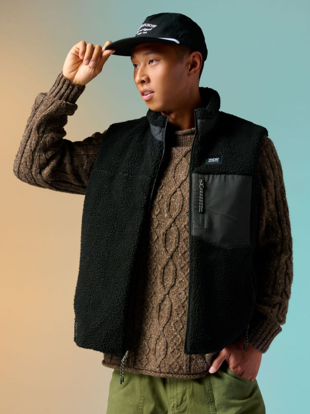 A man in a fisherman’s sweater, fleece vest, and 5-panel hat.